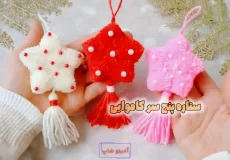 سه عدد آویز ستاره کاموایی پنج پر به رنگ‌های قرمز، سفید و صورتی با بافت مخملی، تزیین مروارید و منگوله آویز در دست.