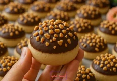 شیرینی خانگی گرد با روکش شکلات قهوه‌ای و تزیین خرده‌های بادام زمینی در دست